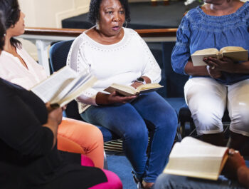 A group of black and African-American woman participating in bible study at church. The main focus is on the senior woman in the middle, talking with a serious expression. She is in her 60s.