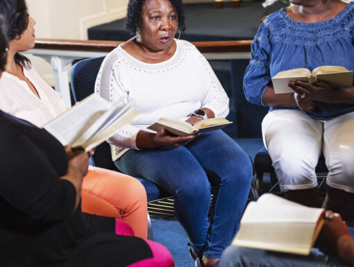 A group of black and African-American woman participating in bible study at church. The main focus is on the senior woman in the middle, talking with a serious expression. She is in her 60s.