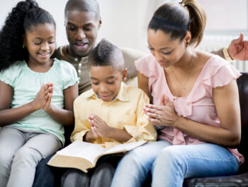 An African-American family sitting on their sofa together, folding their hands for prayer with the bible open.